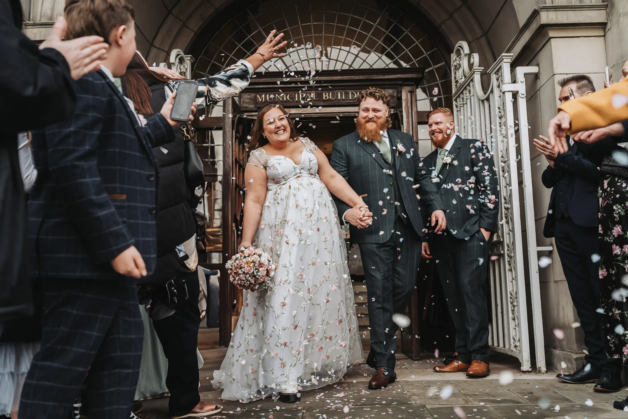 Bride and groom holding hands and beaming under a shower of confetti as they step out of an arched wrought-iron doorway between cheering guests