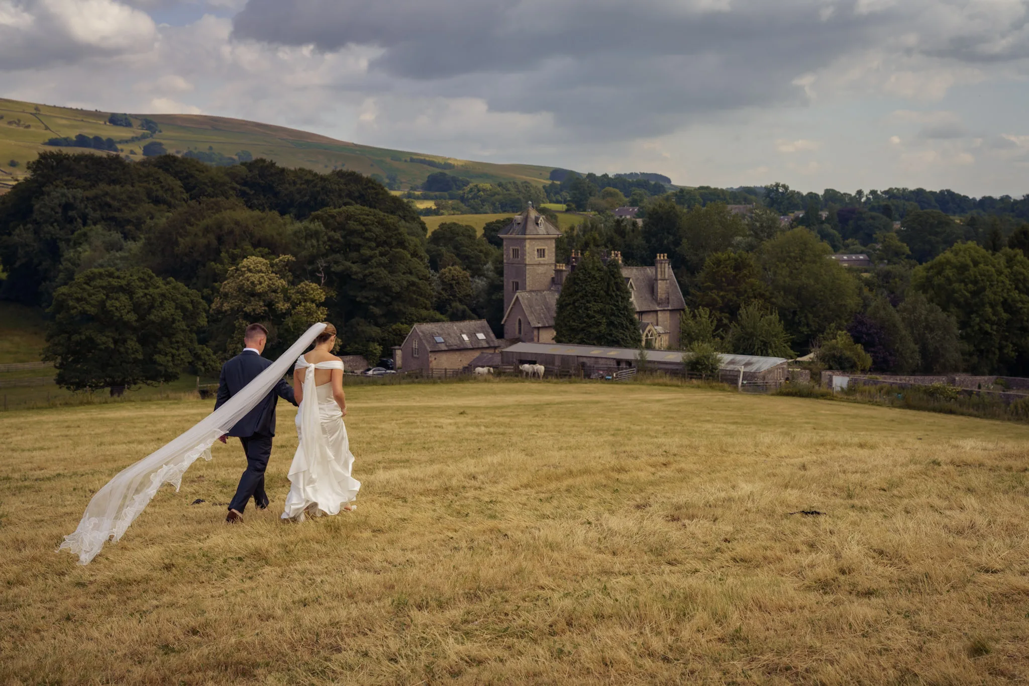 Bride and groom walking across a hay meadow with her veil flying in the wind, a stone country house and the fells behind them